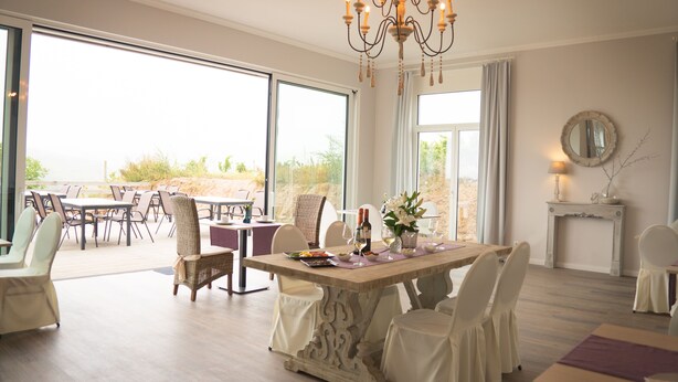 A dining area with a large table, chairs, and a chandelier. There are large windows and a view of the outdoors. - Trautwein - Das Winzerhotel am La Roche (Flonheim)
