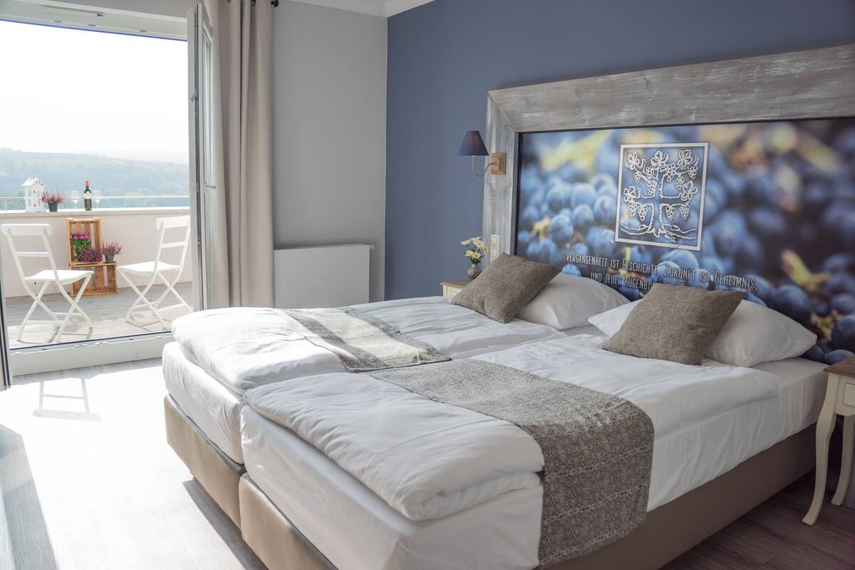 Cama doble con sábanas blancas, una manta gris y dos almohadas. Un cuadro grande enmarca una vid de uvas en la pared.
