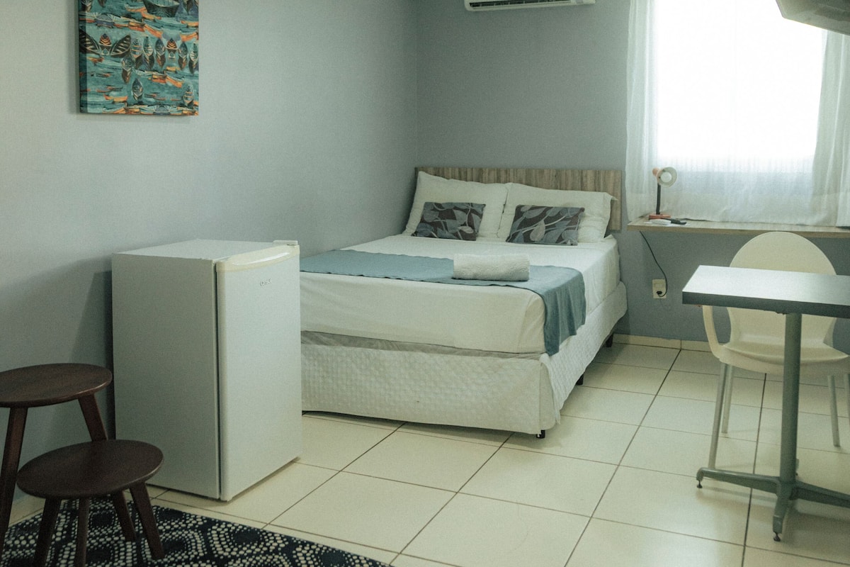 A small, well-lit room with a bed, a desk, a chair, a refrigerator, and a patterned rug.