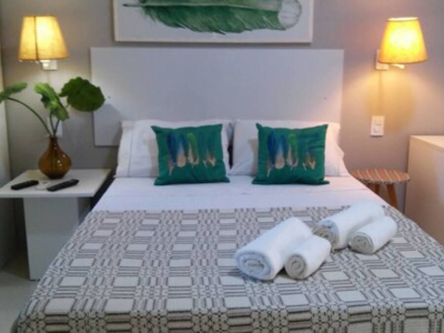 Habitación de hotel con cama, dos almohadas, mesita de noche, lámpara, planta y toallas enrolladas.
