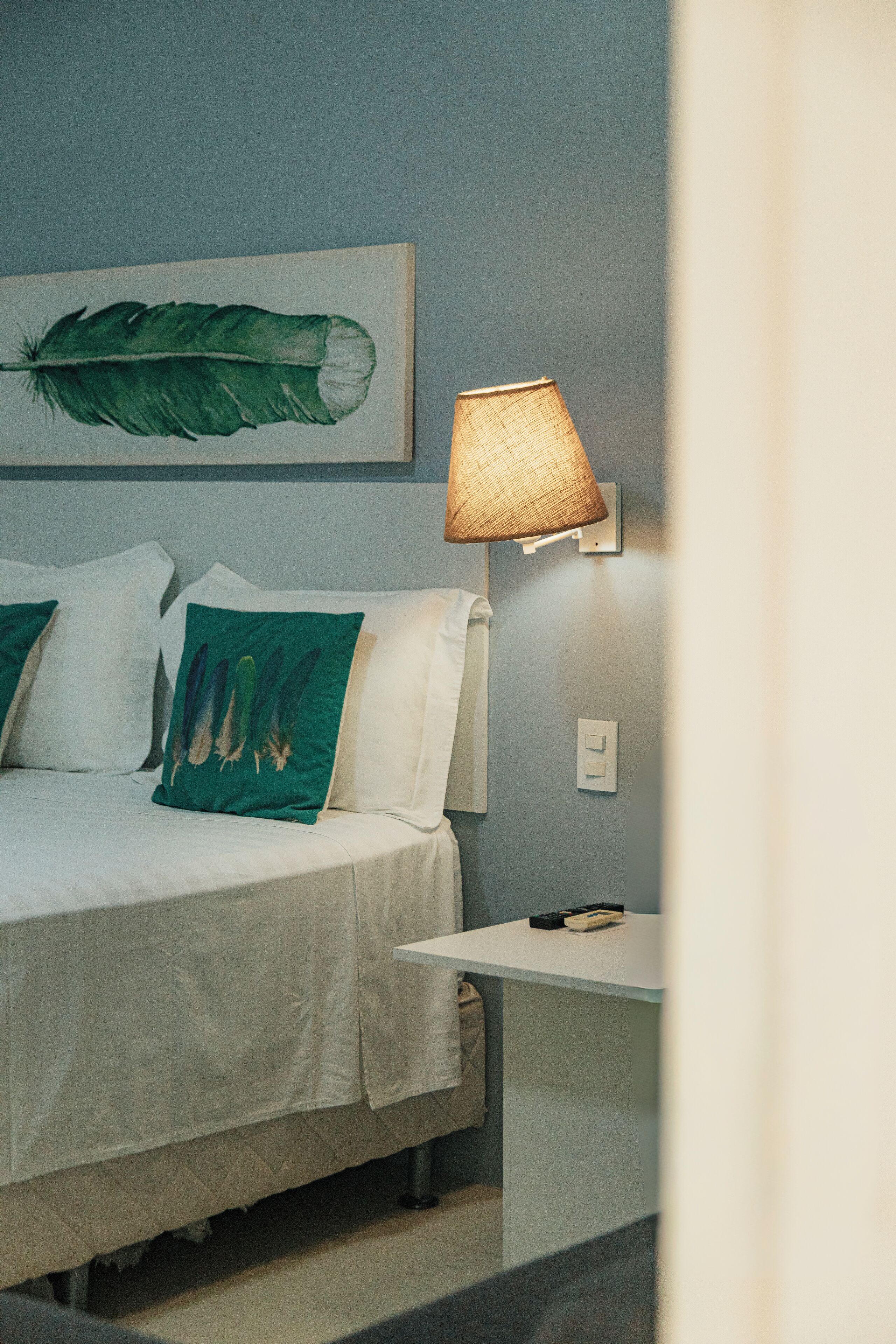 A neatly made bed with white bedding and green pillows, a wall-mounted lamp, and a framed artwork above the bed.