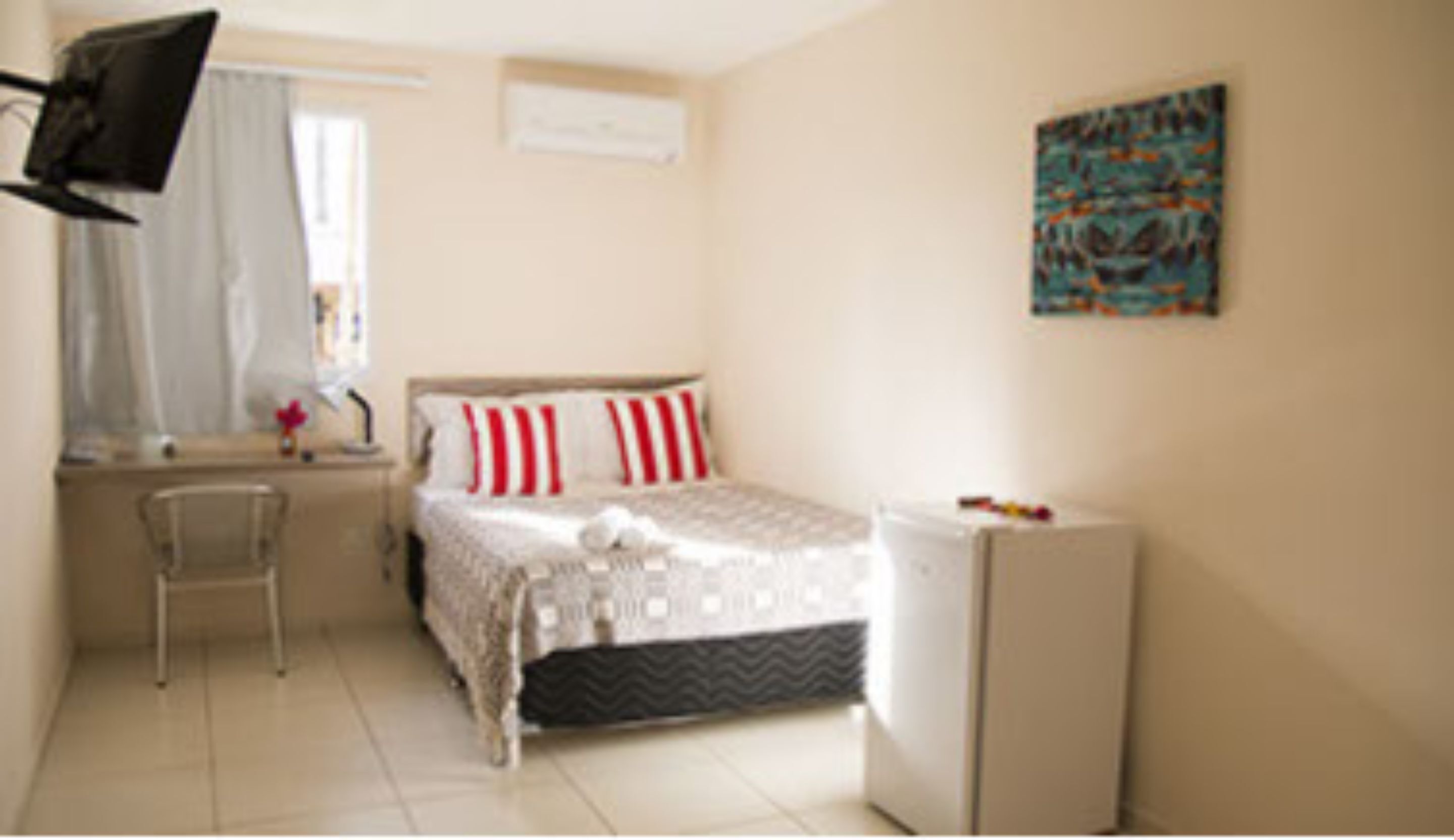 A bedroom with a bed, a desk, a chair, a small refrigerator, and a window with curtains.