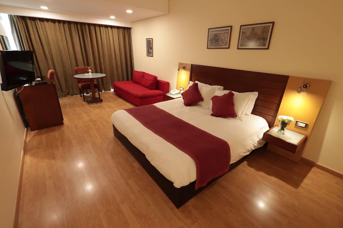Habitación de hotel con una cama grande, un sofá rojo, una mesita pequeña, un televisor y dos lámparas fijadas a la pared.
