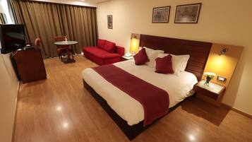 Habitación de hotel con una cama grande, un sofá rojo, una mesita pequeña, un televisor y dos lámparas fijadas a la pared.
