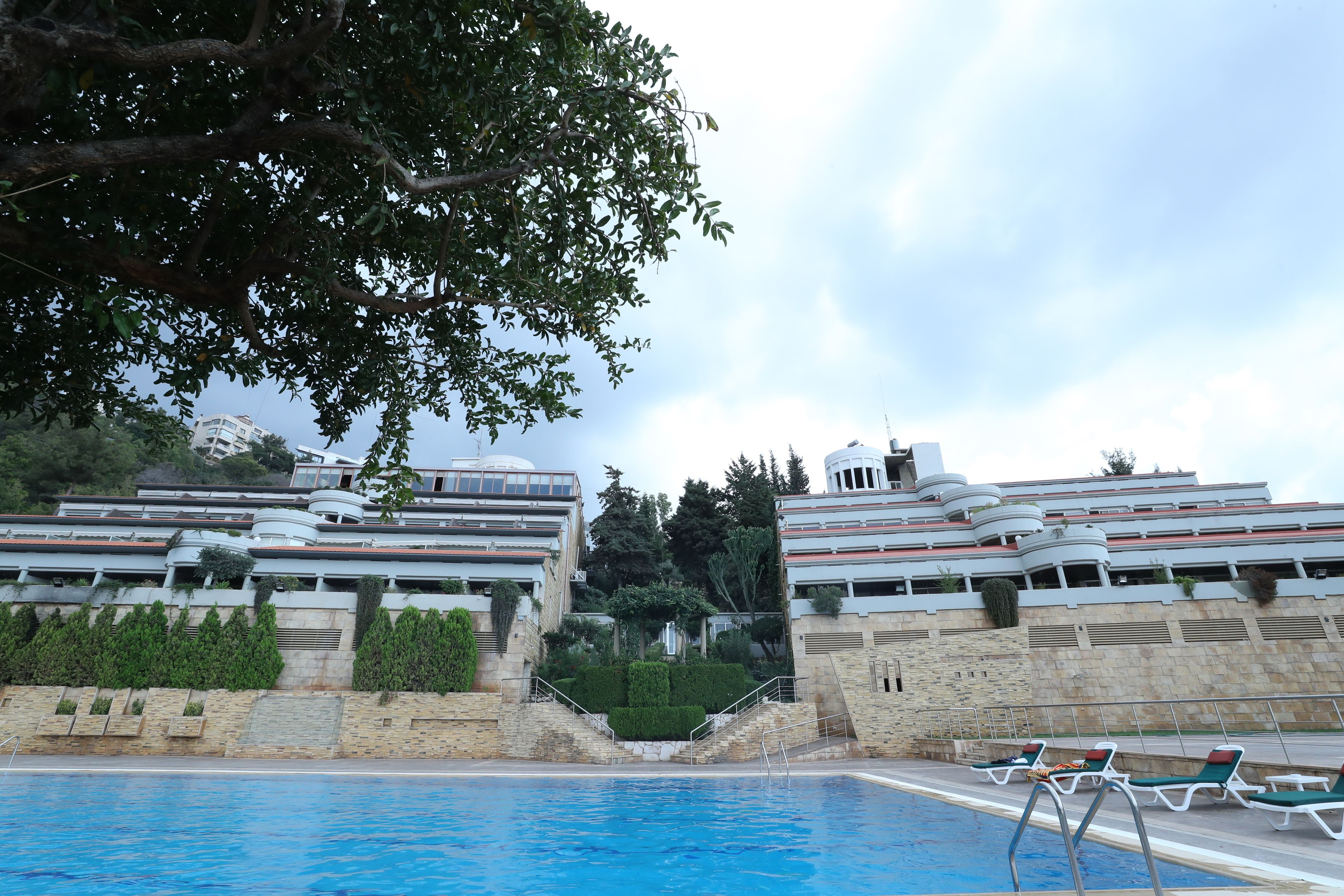 Una piscina con sillones reclinables, una escalera de piedra y edificios modernos al fondo.