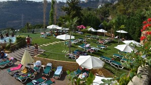 A poolside area with lounge chairs and umbrellas, surrounded by greenery and a view of a cityscape. - Monteverde Hotel (Beit Mery)