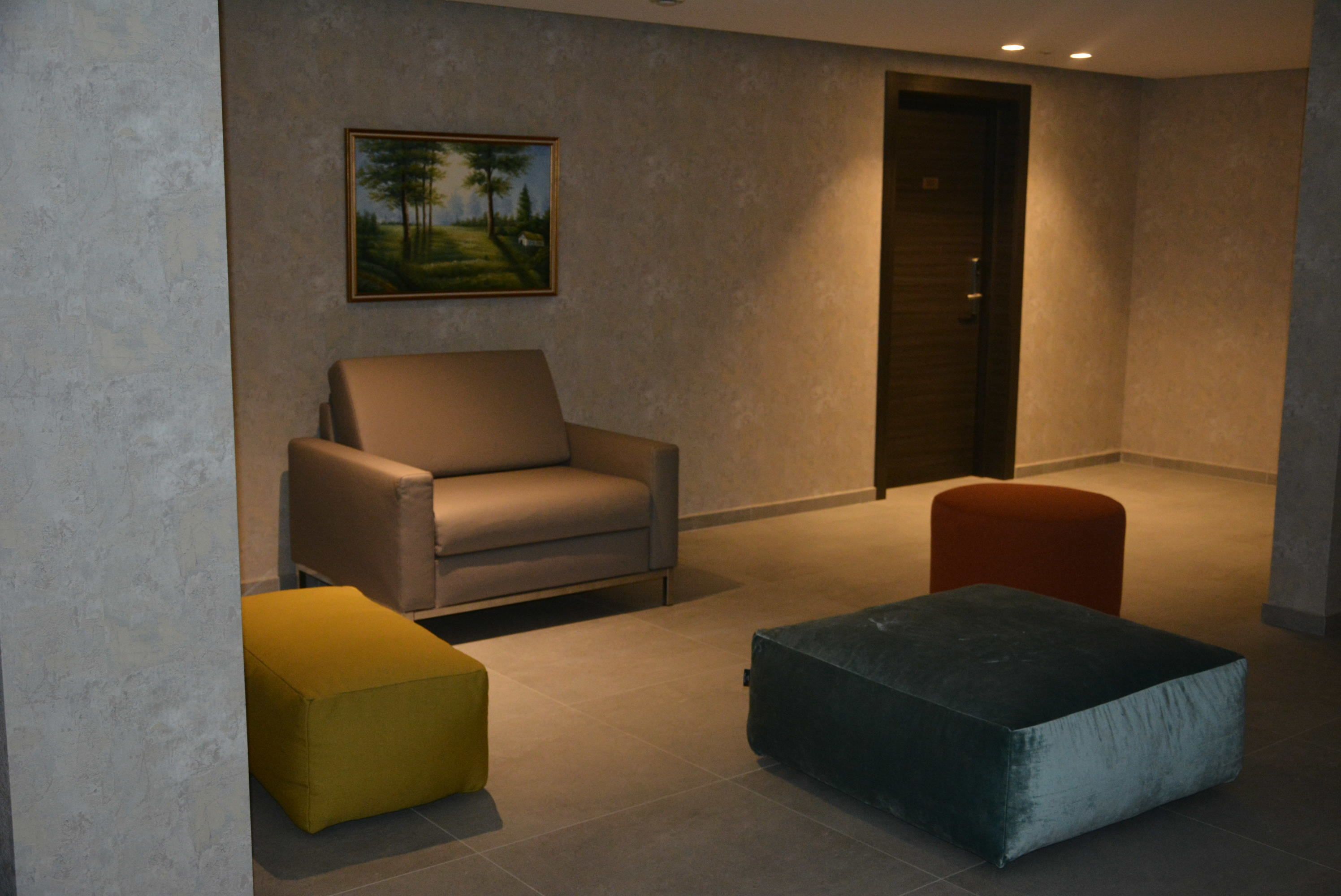 A modern room with a beige sofa, a yellow ottoman, and a dark ottoman. A painting of trees hangs on the wall.