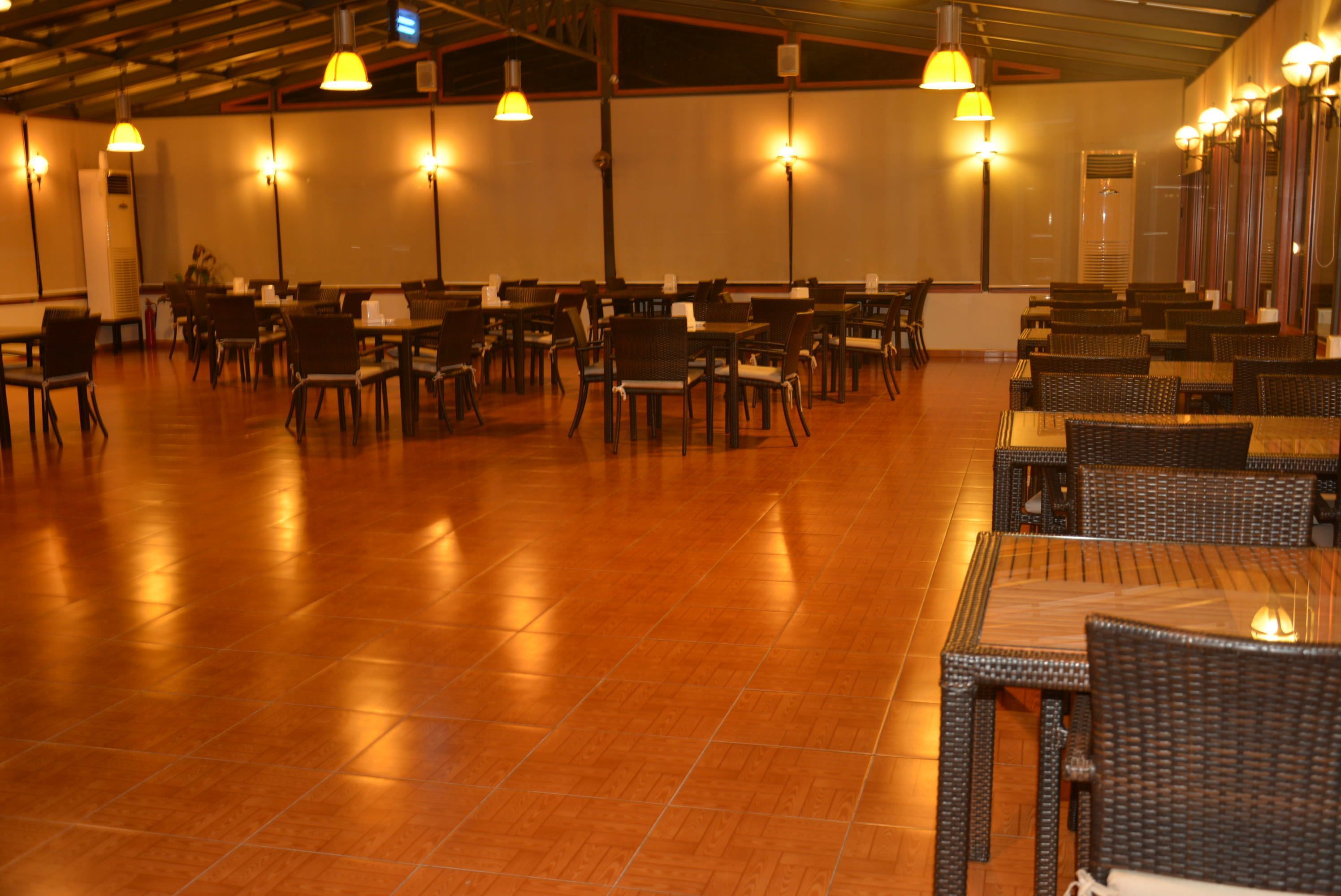 A spacious indoor dining area with multiple tables and chairs, high ceiling with exposed beams, and wall-mounted lighting fixtures.