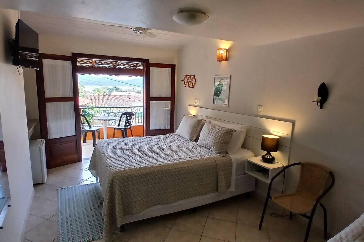 A bedroom with a bed, bedside table, lamp, chair, and a balcony with chairs.