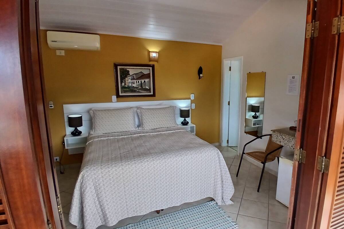 A bedroom with a bed, bedside lamps, a desk with a chair, a wall-mounted light, and a framed picture.