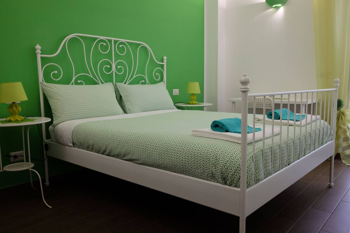 Una cama con estructura metálica blanca y ropa de cama estampada en verde, dos mesitas de noche con lámparas, y una pared verde.