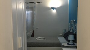 Een kleine, compacte kamer met een bed, een tafel en een stoel. Er is een raam met gordijnen, een wandairconditioner en een lamp op de tafel.