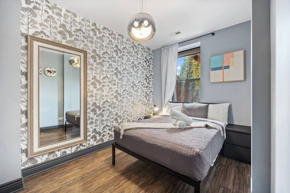Una camera da letto con un grande specchio, un letto con testiera, un comodino e una finestra vista alberi.