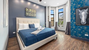 Une chambre à coucher avec un grand lit, une tête de lit, une œuvre d’art accrochée au mur et une fenêtre avec des rideaux.