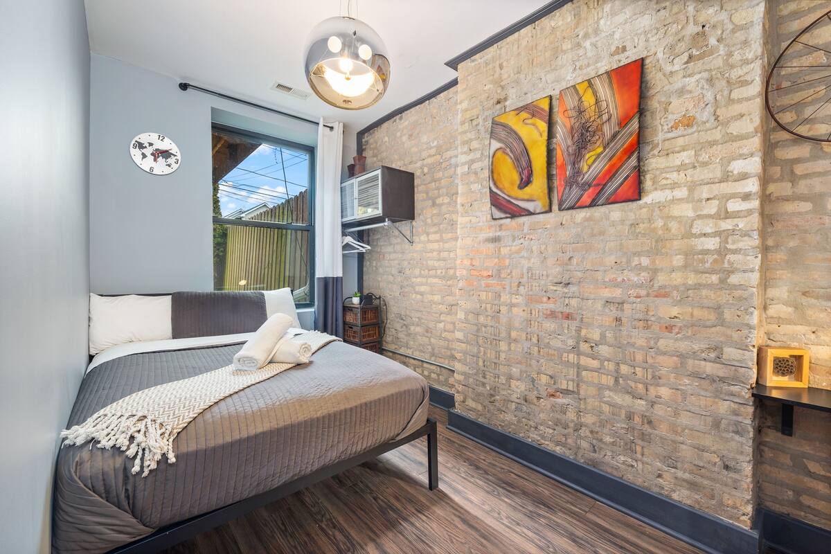 Una camera da letto moderna con una parete in mattoni, un letto con copripiumino grigio, una sveglia mondiale e un quadro.