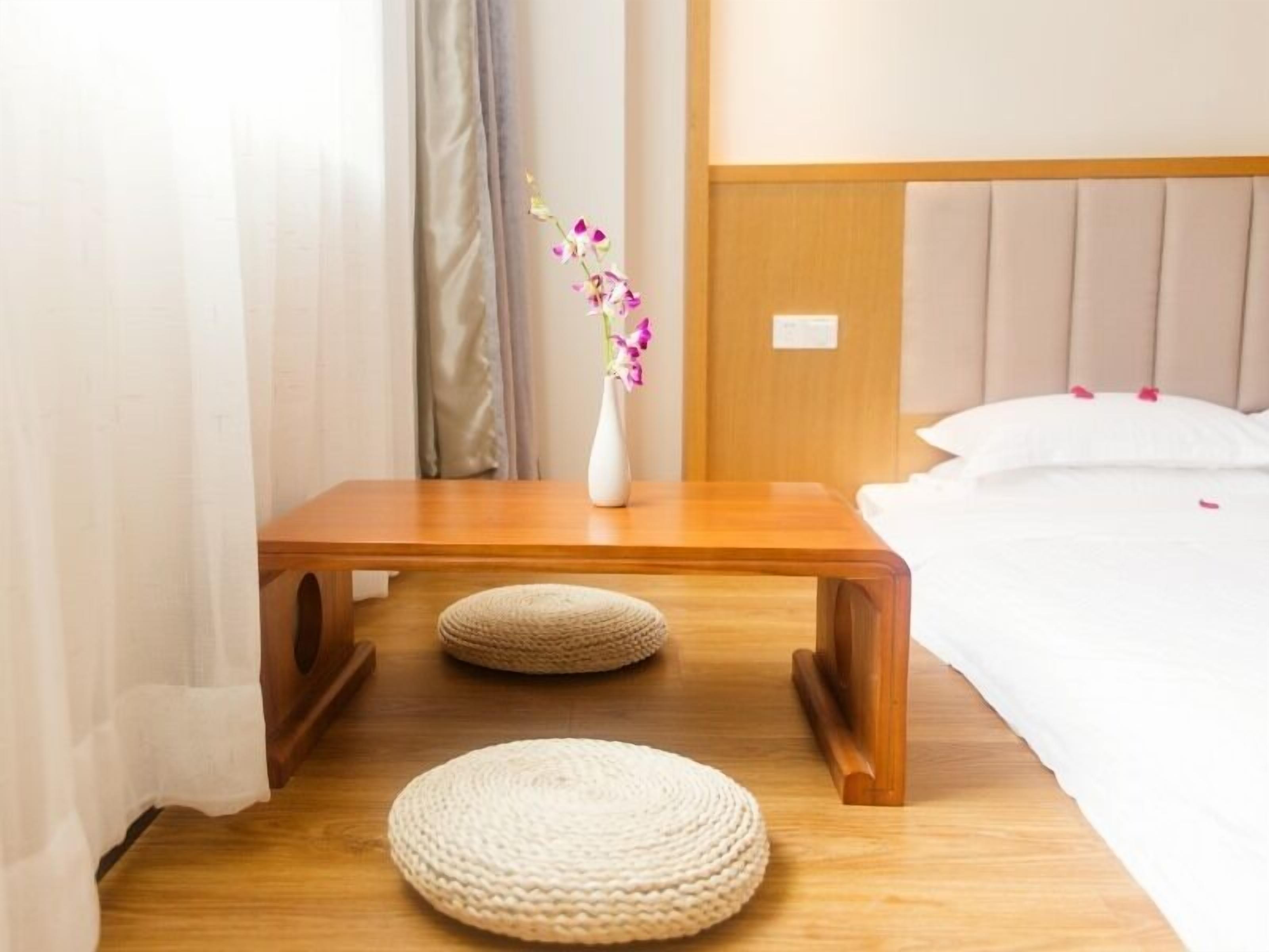 A neatly arranged hotel room with a wooden nightstand, a white vase with flowers, a round woven stool, and a bed with white bedding.