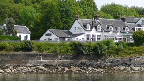 Un grand bâtiment blanc, doté de nombreuses cheminées, qui ressemble probablement à un hôtel, situé au bord d’une côte rocheuse, avec une végétation luxuriante en arrière-plan.