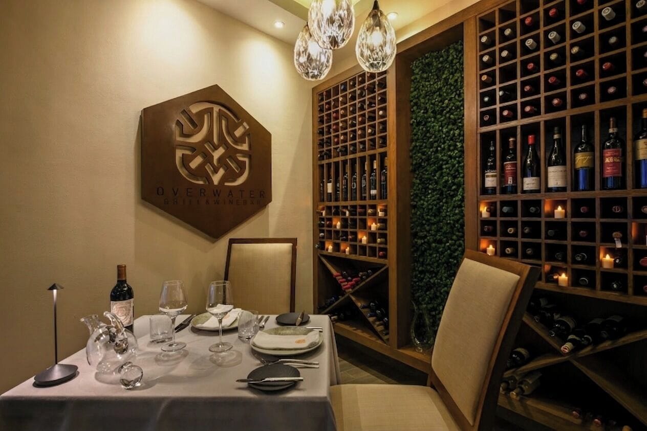 A dining table set with wine glasses, plates, and cutlery, a wine rack with bottles, and a logo on the wall.