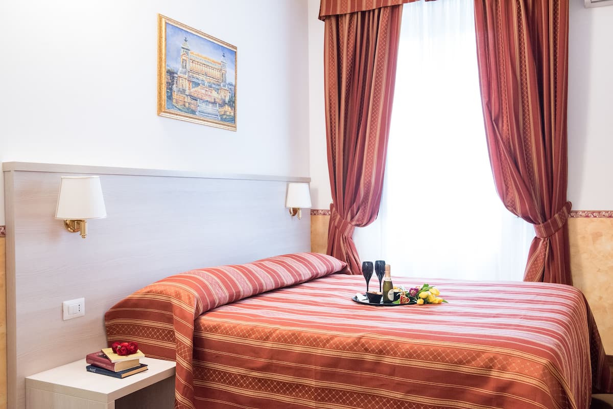 A hotel room with a bed, a nightstand with books and a bottle, a painting on the wall, and a window with curtains.