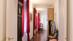 Um corredor com piso de madeira, cortinas vermelhas, um relógio, uma cadeira e um porta-roupas.