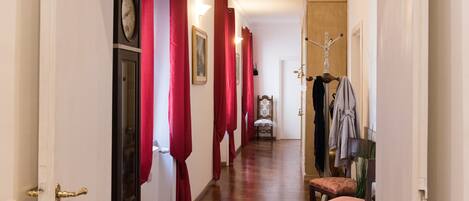 A hallway with a wooden floor, red curtains, a clock, a chair, and a coat rack.