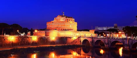 A historic building with a large central tower, a bridge with three arches, and a calm river reflecting the structure.