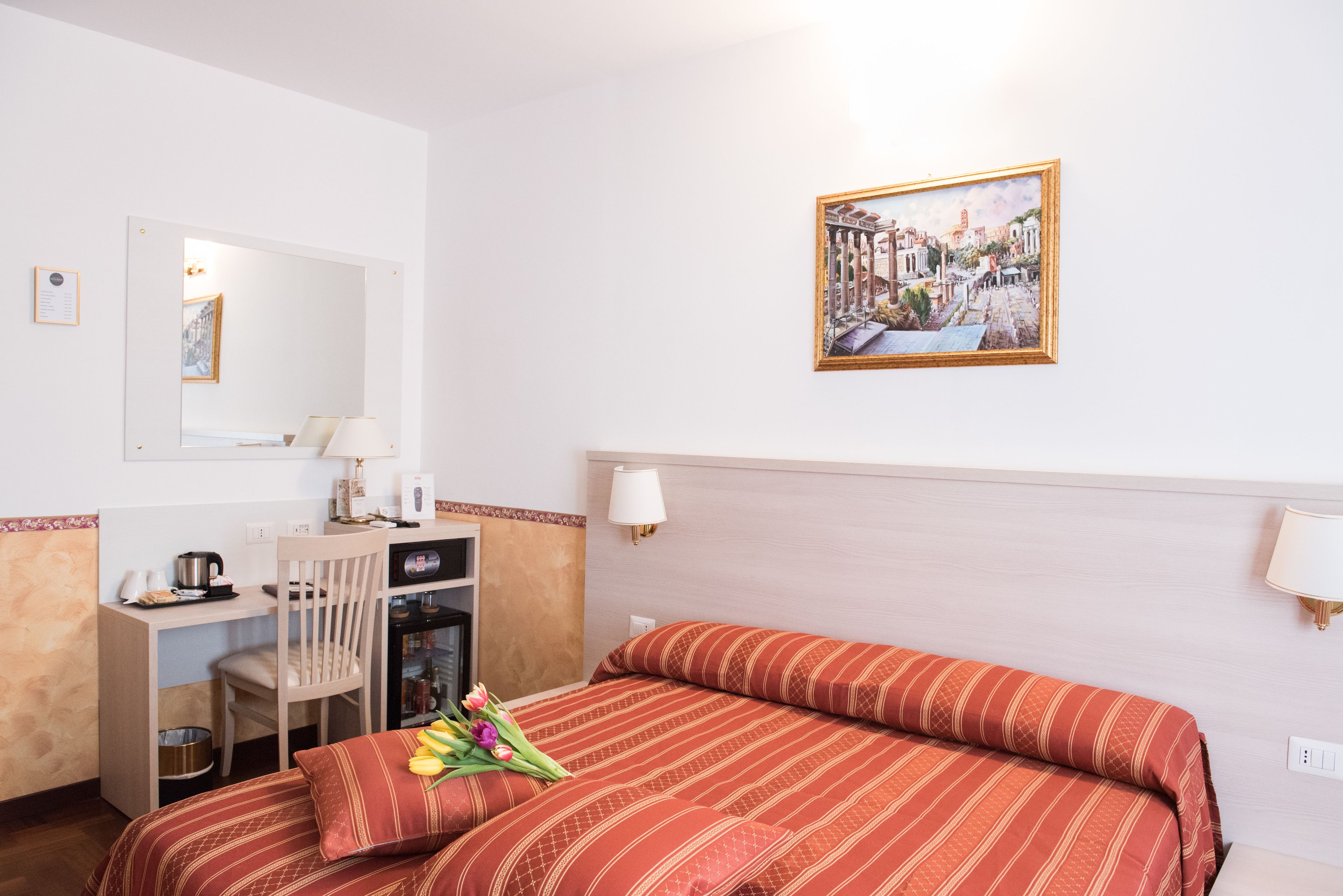 A hotel room with a bed, a desk with a chair, a microwave, a kettle, and a small refrigerator. There is a painting on the wall and a window with curtains.