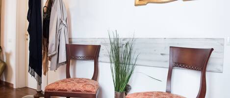 Dos sillas de madera con cojines estampados, un florero con plantas verdes y un espejo con marco dorado sobre una pared blanca.