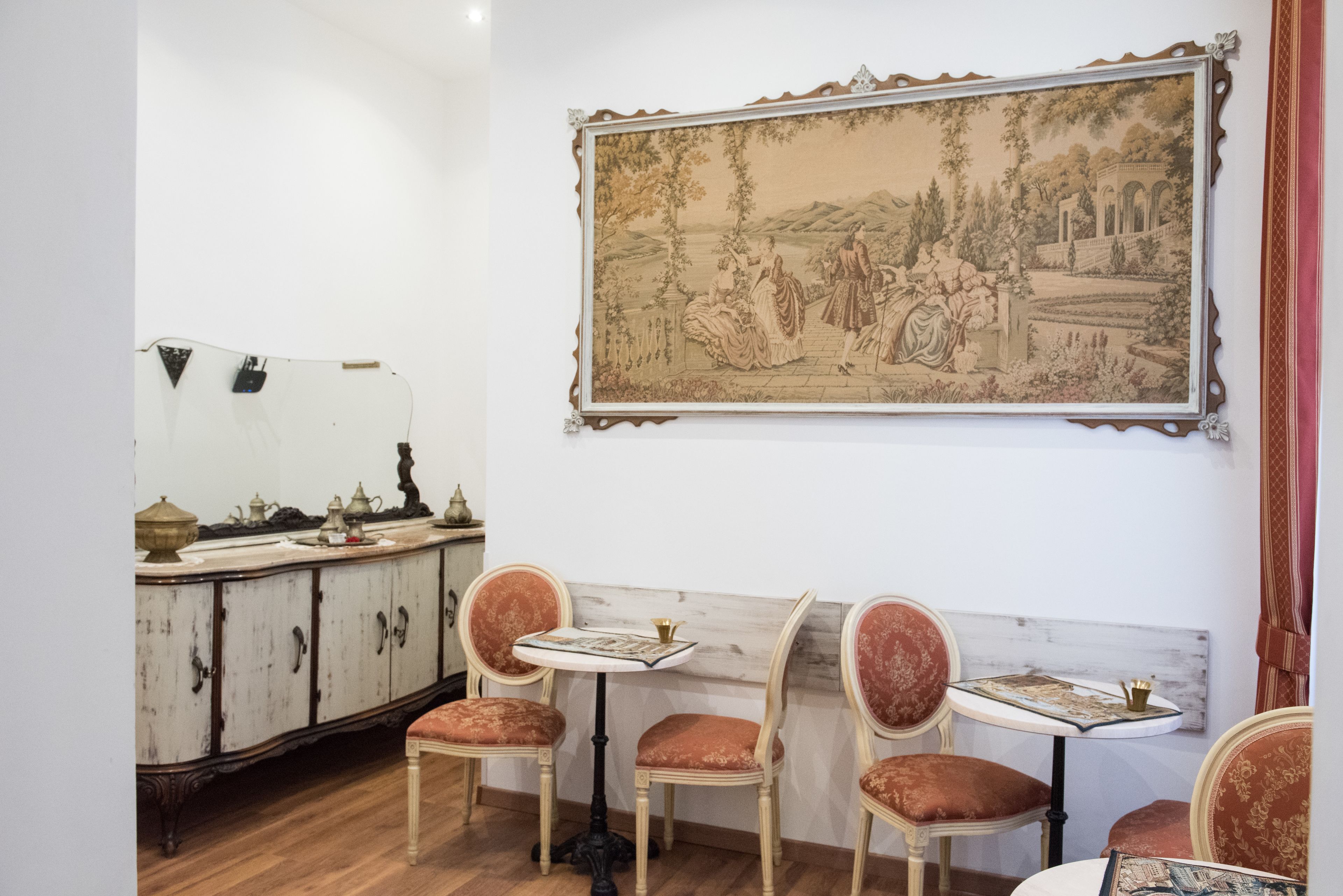 Et spiseområde med et bord og stole i vintage-stil, et stort indrammet landskabsmaleri og et træskab med dekorative genstande.