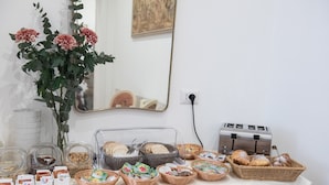 Mesa de buffet com diversos alimentos, incluindo pão, doces e acompanhamentos, dispostos sobre uma toalha de mesa branca.