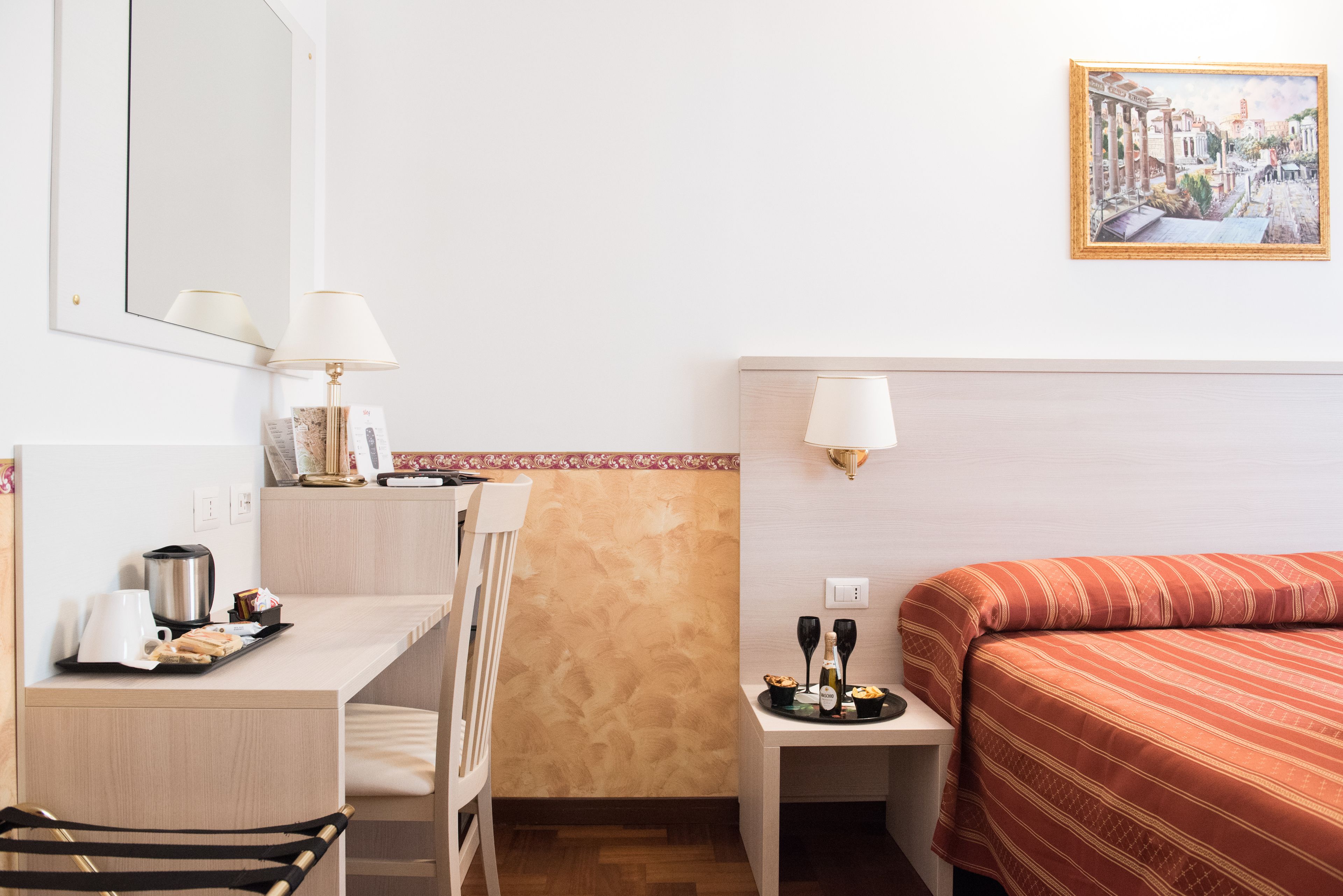 Habitación de hotel con cama, escritorio, silla, una mesita con lámpara, un cuadro en la pared y una luz en la pared.