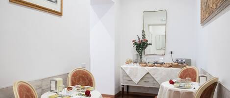 A dining area with two round tables, each with a white tablecloth and floral chairs. A mirror hangs on the wall, and a painting is visible above the mirror.