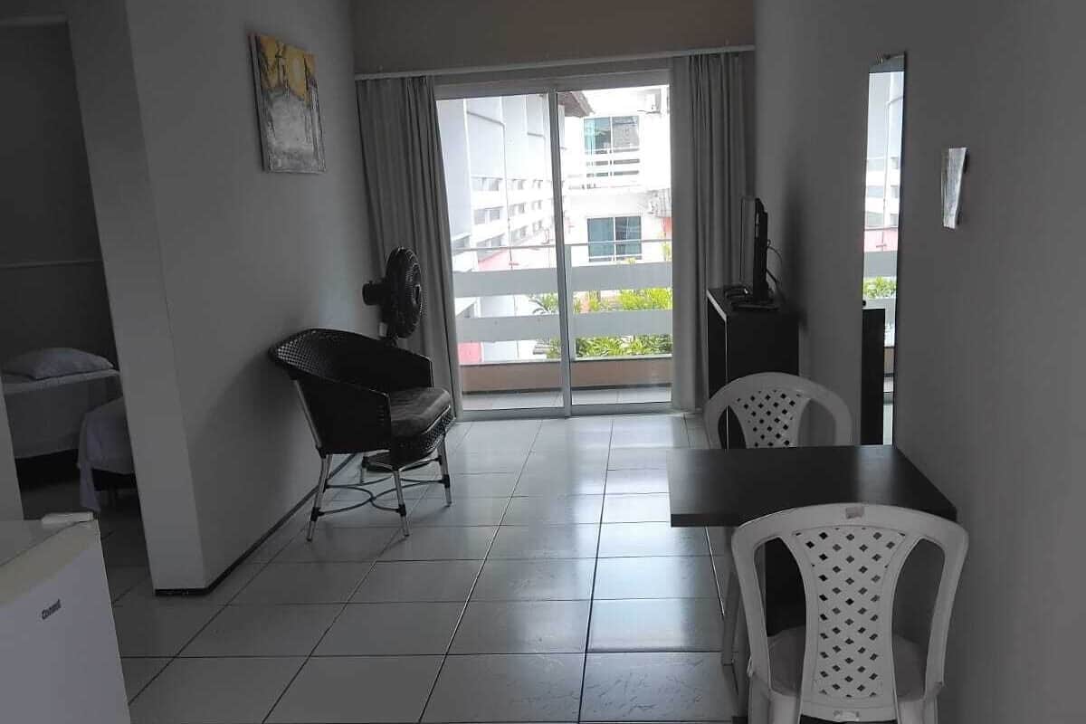 A small, modern room with a dining table, two chairs, a refrigerator, a black chair, and a sliding glass door leading to a balcony.