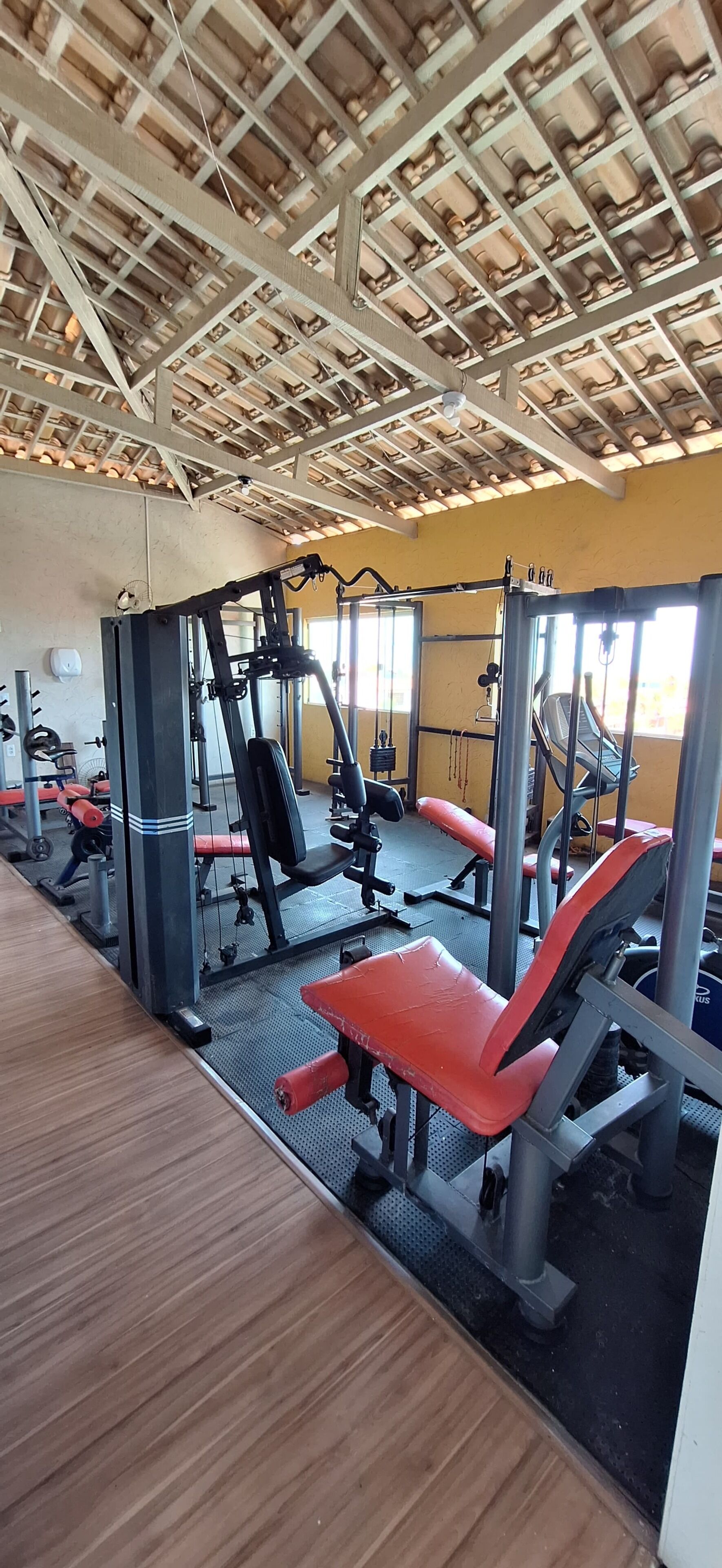 A gym with various exercise machines, wooden flooring, and a high ceiling with exposed beams.