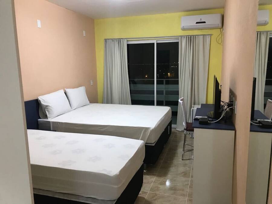 Quarto de hotel com duas camas, uma escrivaninha, uma cadeira e uma janela grande com cortinas.