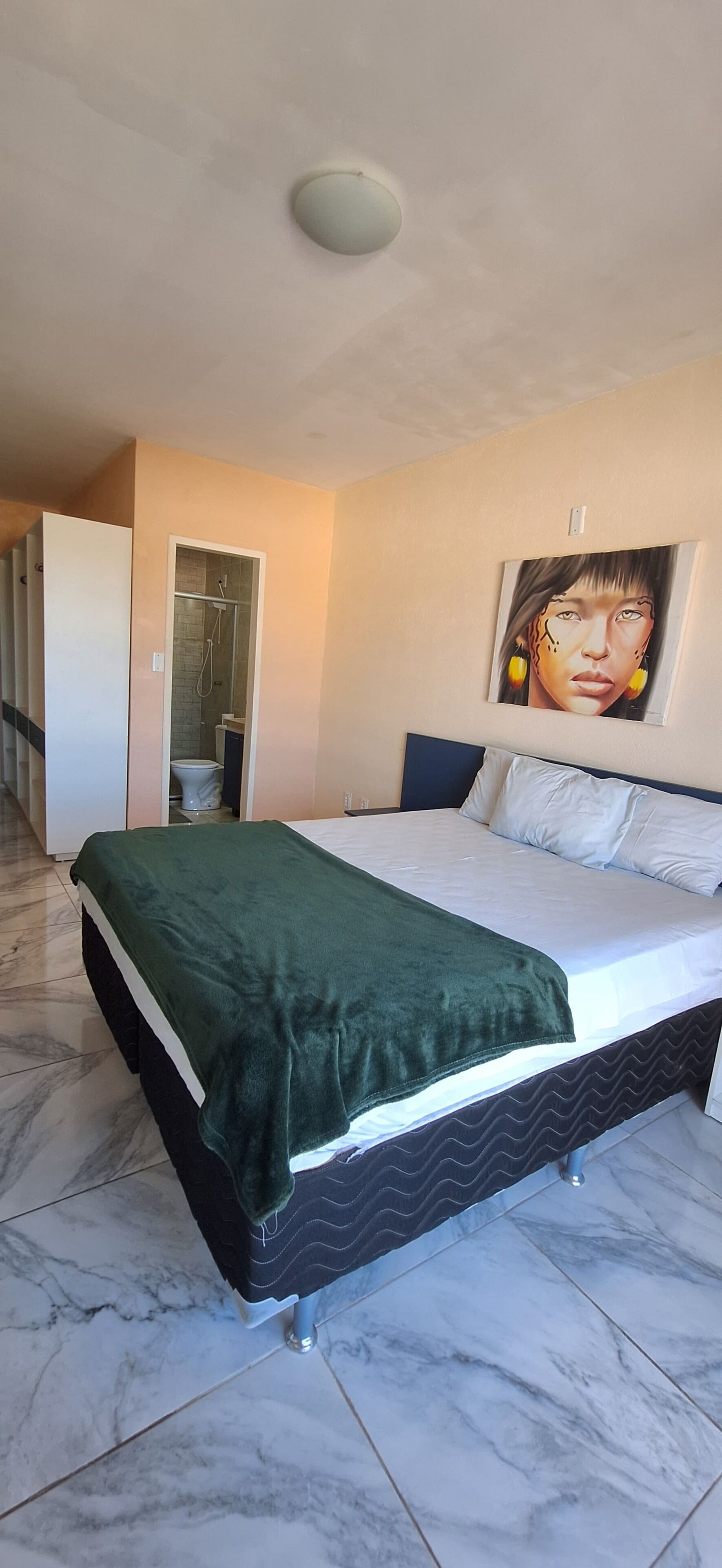 Un dormitorio con cama, manta verde, un cuadro en la pared y un baño con ducha.