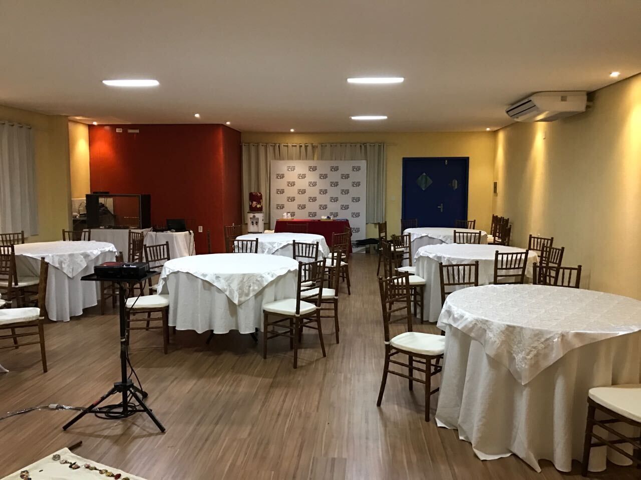 Un área de comedor con varias mesas redondas cubiertas con manteles blancos, sillas de madera y un escenario con fondo rojo.