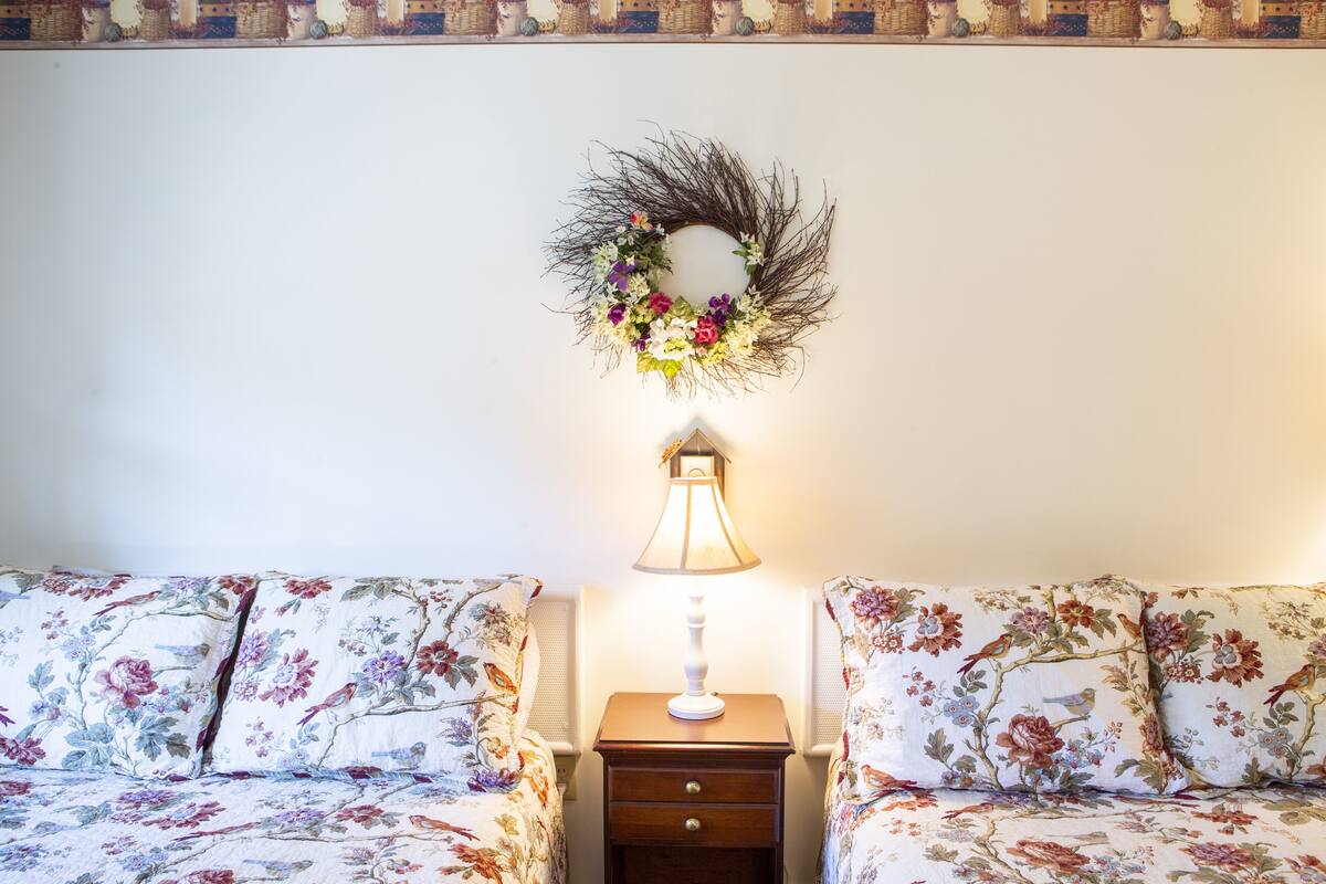 A bedroom with two floral-patterned beds, a nightstand with a lamp, and a decorative wreath on the wall.