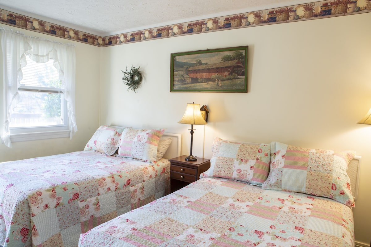 A bedroom with two beds, a nightstand, a lamp, a painting, and a window with curtains.
