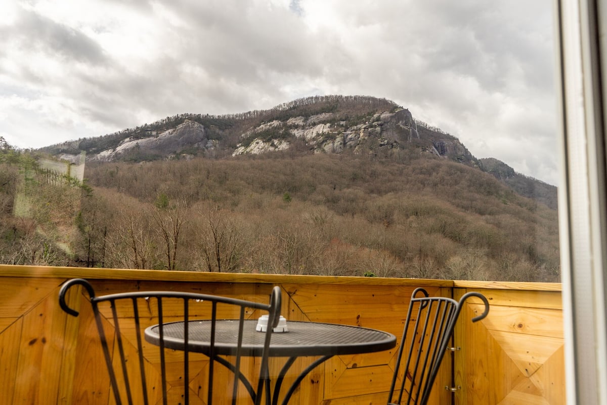 A wooden deck with a table and chairs, overlooking a mountainous landscape with a forested hill.