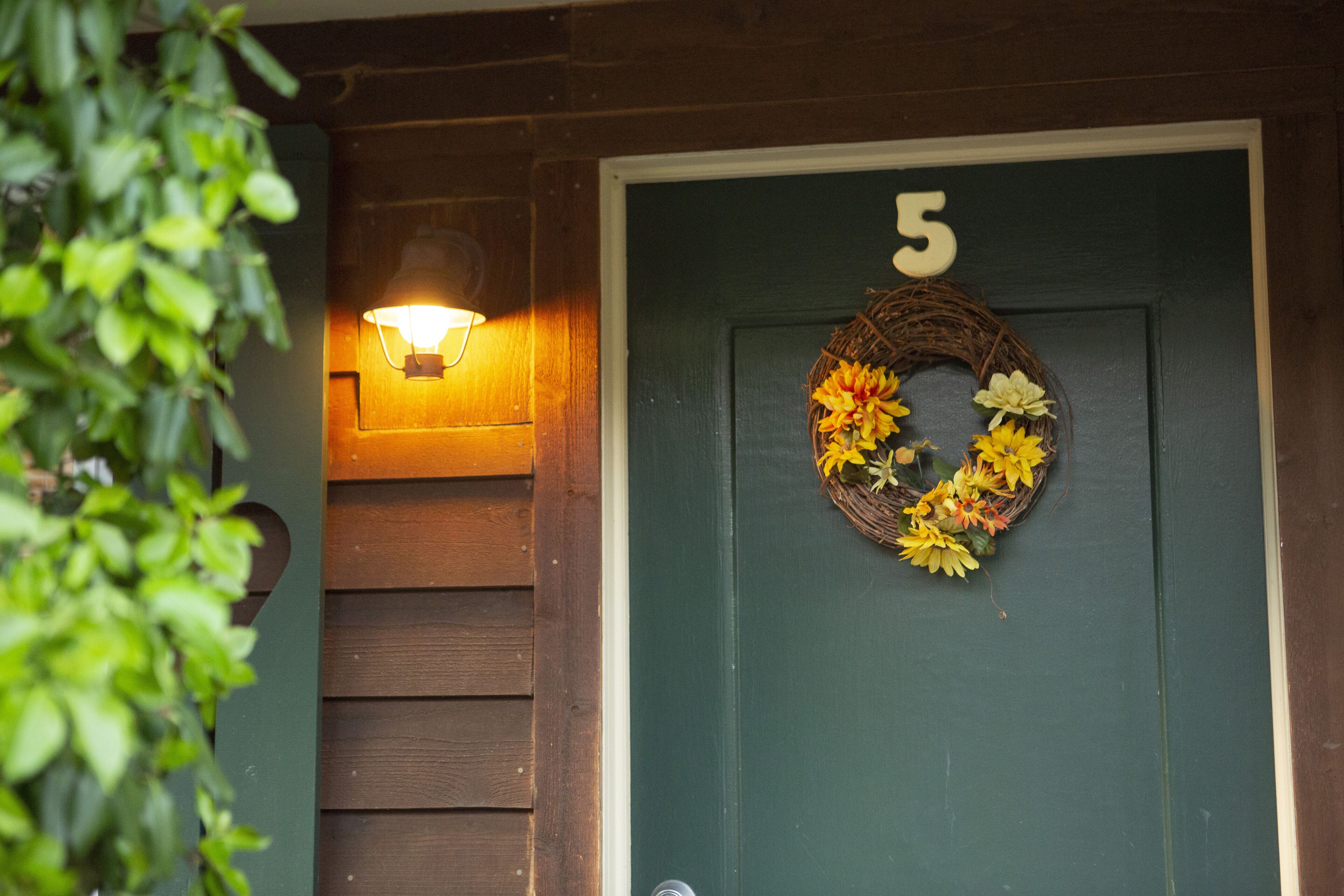 A green door with the number 5, a lit wall-mounted lamp, and a wreath with yellow flowers.