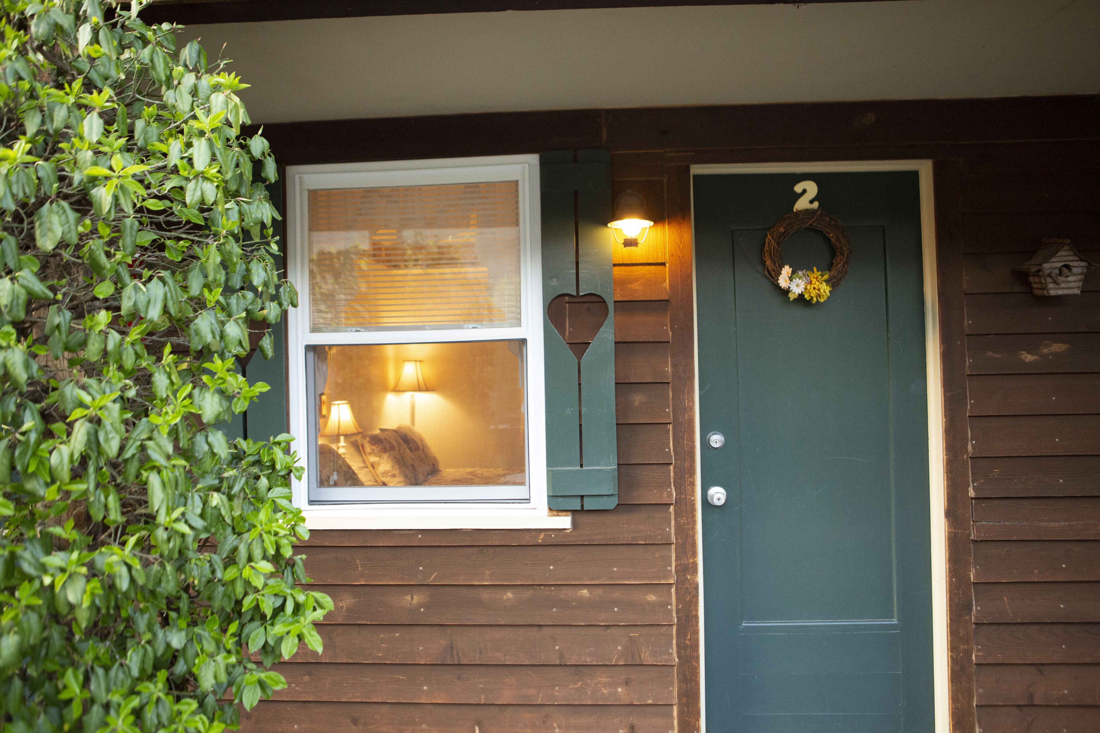 A wooden house with a green door, a window with a lamp inside, and a wreath on the door.