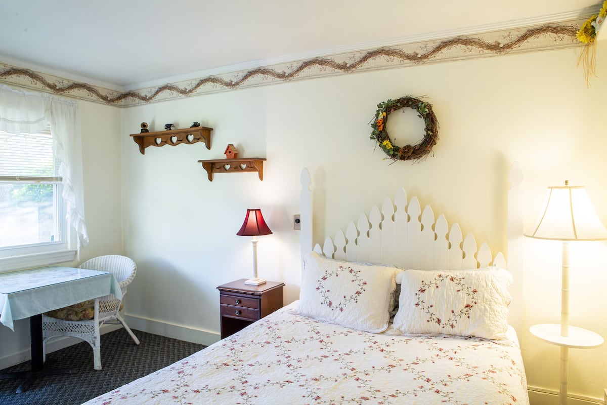 A bedroom with a bed, a desk, a chair, a nightstand, and a lamp.