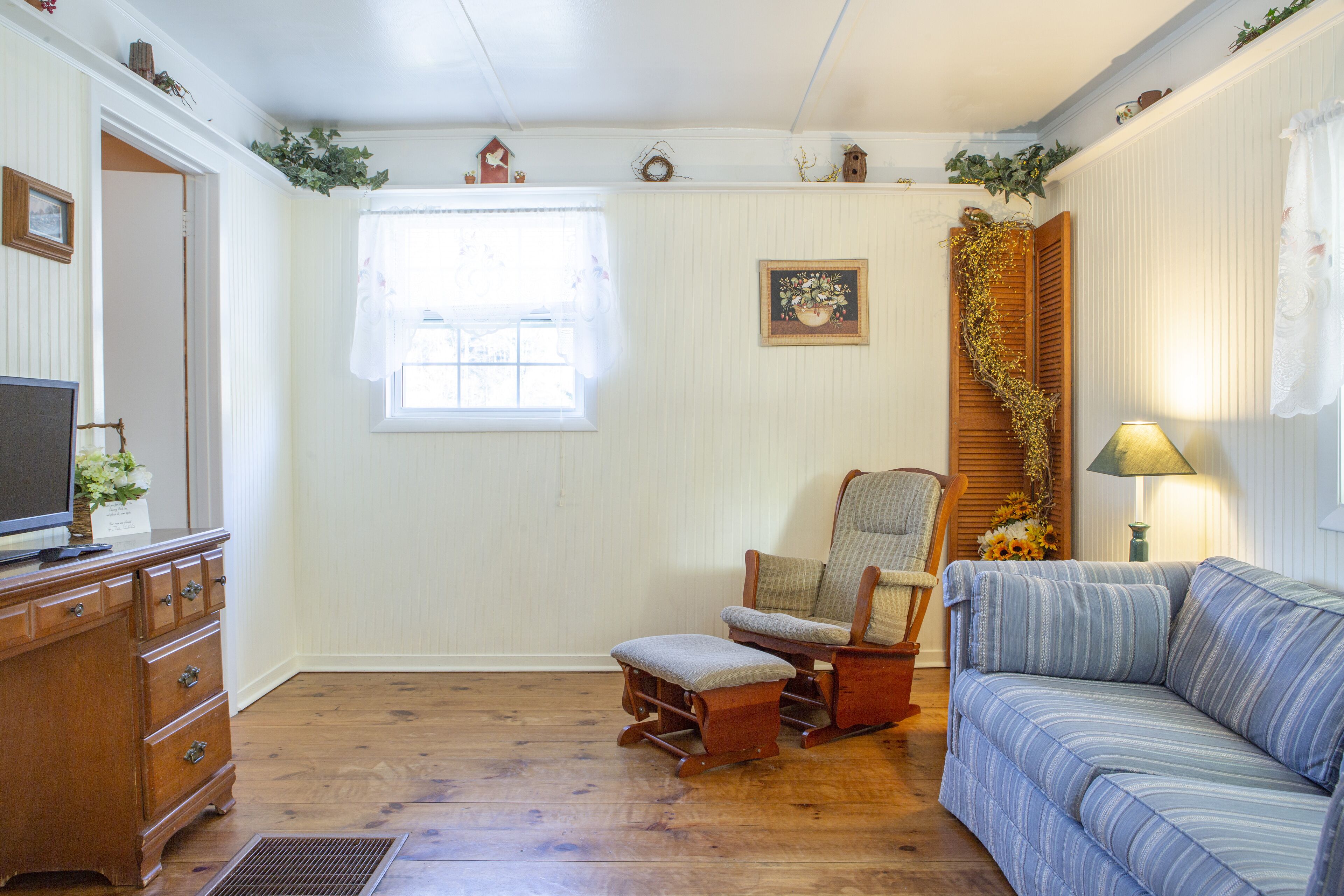 A cozy living room with a wooden floor, a sofa, a rocking chair, a television on a wooden cabinet, and a window with a white curtain.