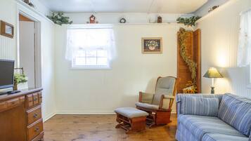 A cozy living room with a wooden floor, a sofa, a rocking chair, a television on a wooden cabinet, and a window with a white curtain.