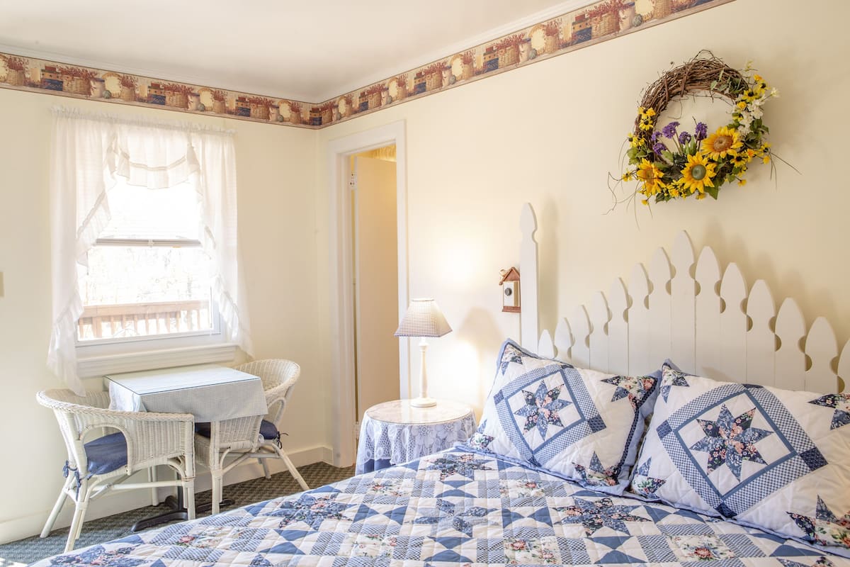 A bedroom with a bed, a small table, two chairs, a lamp, and a decorative wreath on the wall.