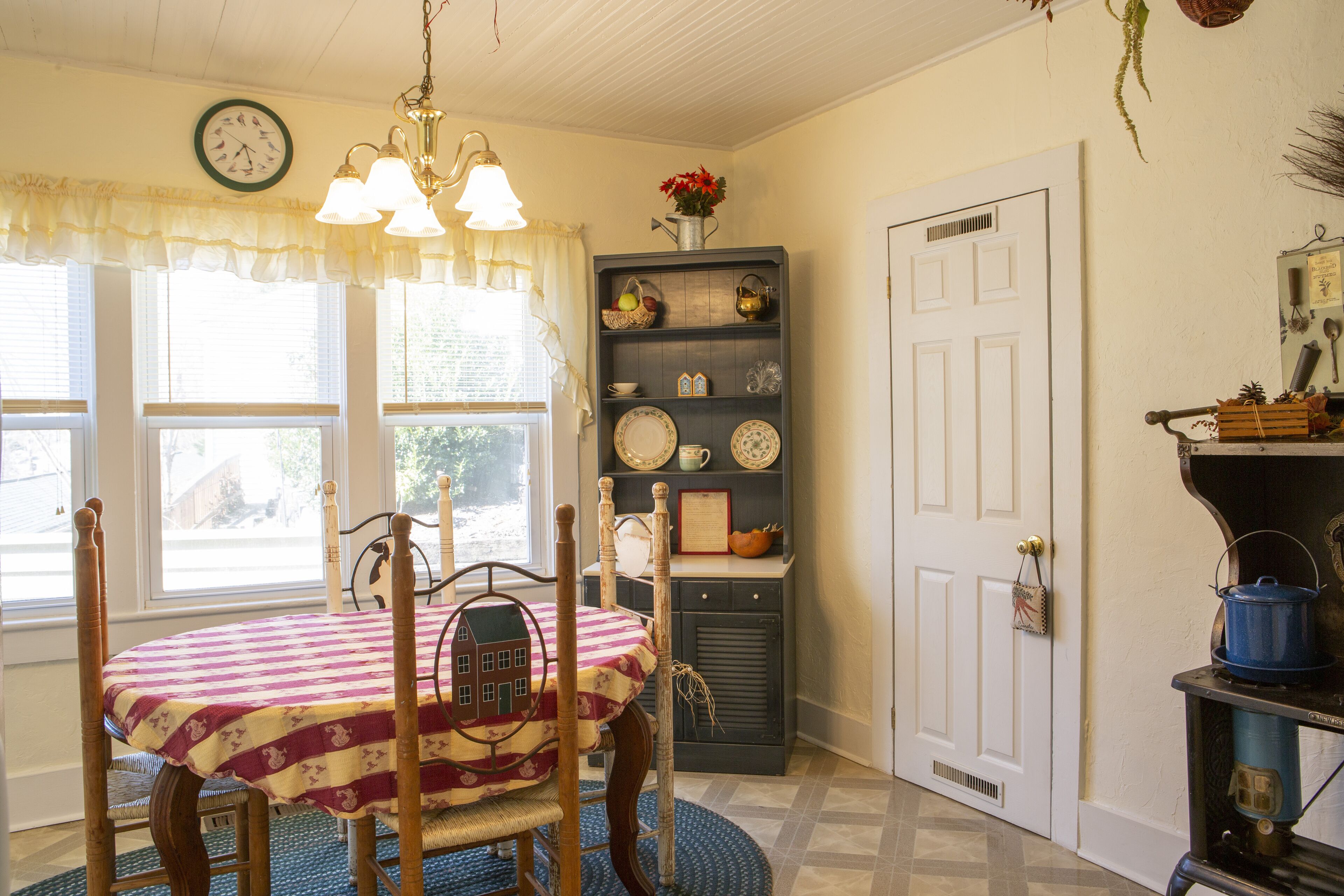 Un coin repas avec une table dressée pour quatre personnes, un lustre, une horloge au mur et une fenêtre avec des rideaux.