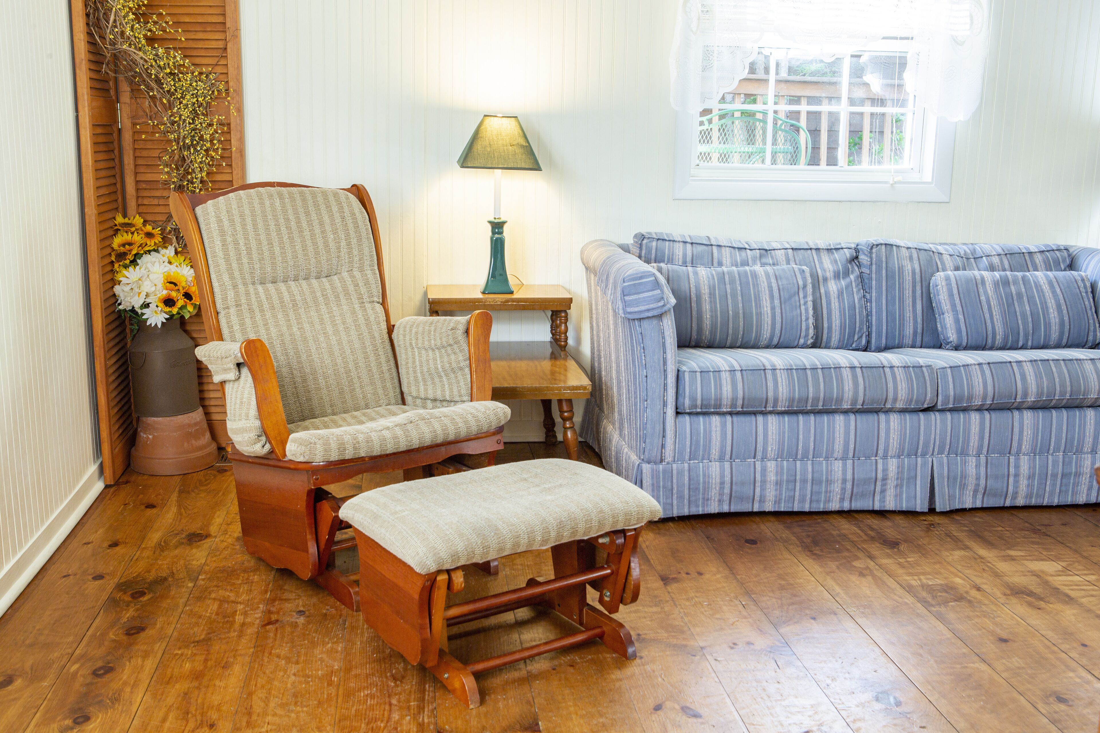 A living room with a wooden floor, a striped sofa, a rocking chair, a small table with a lamp, and a window with a view of buildings.