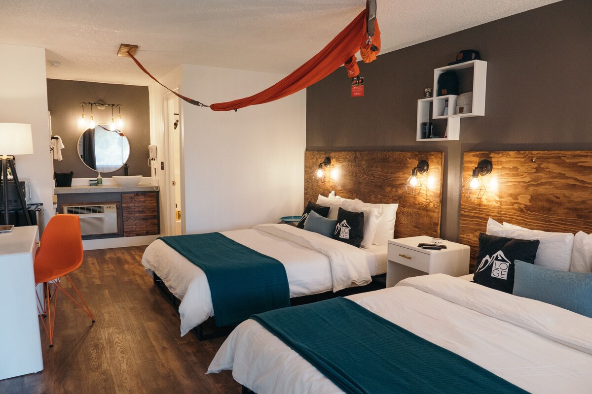 Une chambre d’hôtel avec deux lits, un bureau, une chaise, une coiffeuse avec un miroir et un baldaquin rouge suspendu.