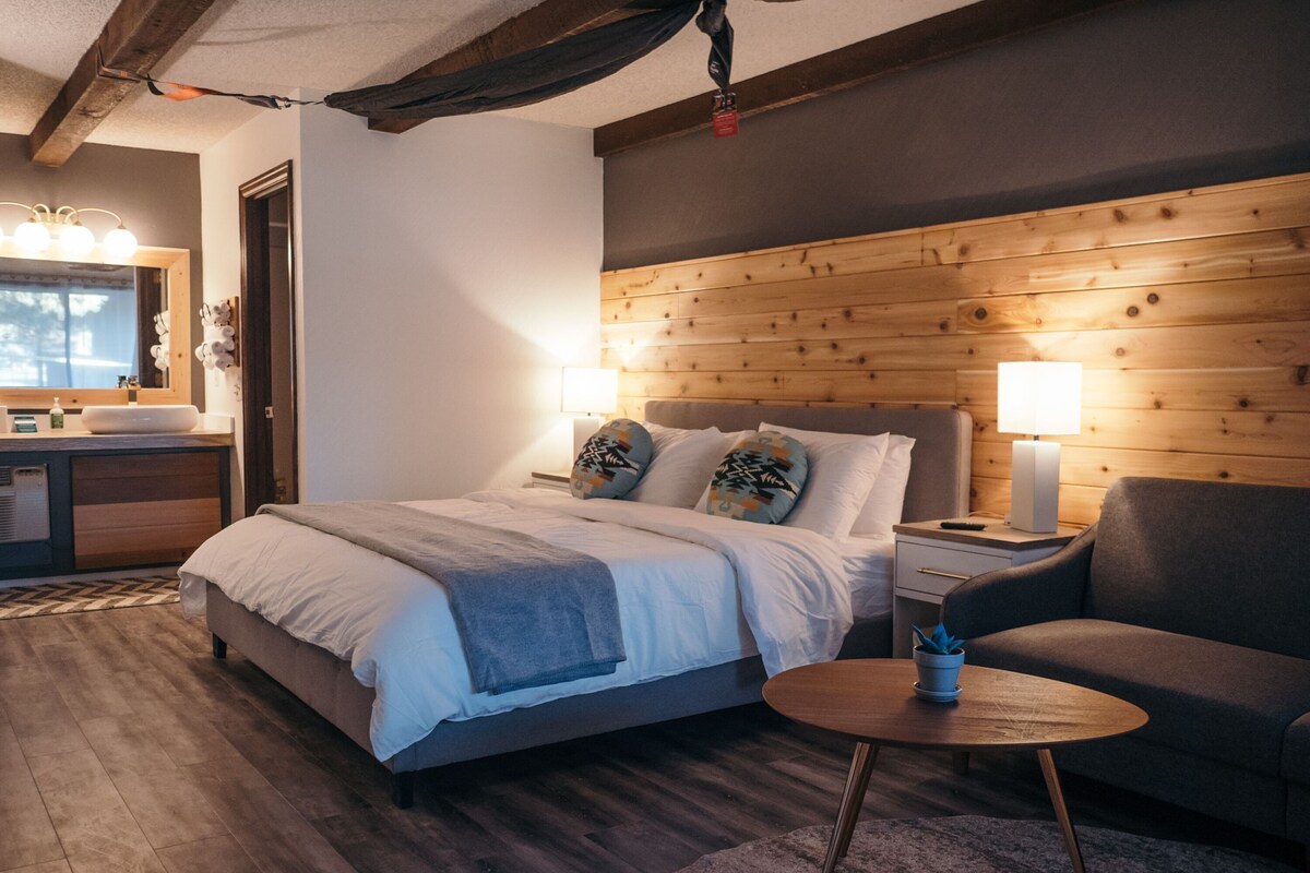 Une chambre à coucher moderne avec une tête de lit en bois, un grand lit avec de nombreux oreillers, une table de chevet avec une lampe, une petite table ronde et un canapé.